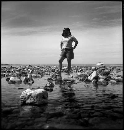 A female figure wearing a gas mask standing on a rocky lake shore with a pleasure boat in the distance.